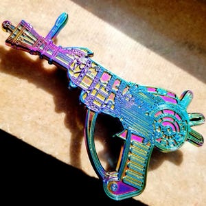 May include: A rainbow iridescent enamel pin of a detailed, futuristic, sci-fi gun with a clock face on the side.