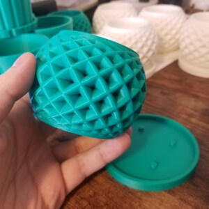 May include: A teal-colored, geometric-patterned planter pot with a matching saucer. The pot has a spherical shape with a grid-like design. Other similar planters are visible in the background. The pot is held by a hand.