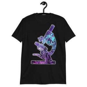 May include: Black t-shirt featuring a detailed illustration of a microscope. The microscope design is filled with a vibrant galaxy pattern in shades of purple, blue, and teal, outlined in white. The shirt is hanging on a black hanger.