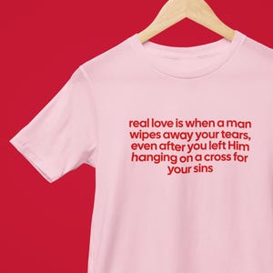 May include: A light pink t-shirt with the text "real love is when a man wipes away your tears, even after you left Him hanging on a cross for your sins" in red. The shirt hangs on a wooden hanger.