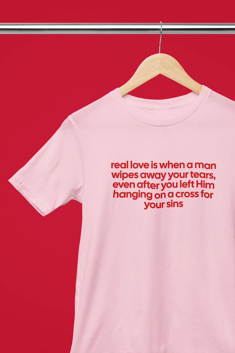 May include: A light pink t-shirt with the text "real love is when a man wipes away your tears, even after you left Him hanging on a cross for your sins" in red. The shirt hangs on a wooden hanger.