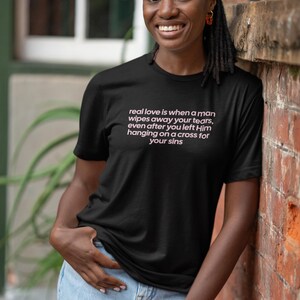May include: Black t-shirt with the text "real love is when a man wipes away your tears, even after you left Him hanging on a cross for your sins" in pink. The shirt is worn by a person in light blue jeans.
