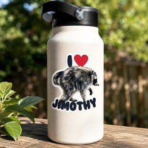 May include: A matte off-white insulated water bottle with a black lid featuring a sticker of a raccoon. The sticker displays the text I, a red heart symbol, and the name JIMOTHY in dark blue capital letters below the raccoon illustration.