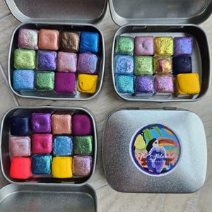 Peut inclure: Quatre petites boîtes métalliques ouvertes remplies de godets de peinture aquarelle carrés et colorés. Les couleurs comprennent des nuances de rose, de violet, de bleu, de vert et de jaune. Un couvercle de boîte présente un autocollant circulaire avec un toucan et le texte "The Alchemist".