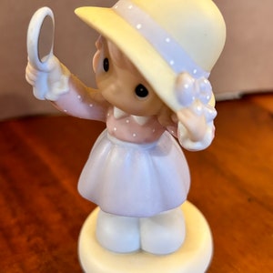 May include: A porcelain figurine of a girl wearing a yellow hat with a blue ribbon and a pink and white polka dot dress. She is holding a small mirror and looking at her reflection.