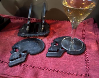 Peut inclure: Deux sous-verres en forme de crâne noirs avec un verre de vin blanc sur l'un des sous-verres. Les sous-verres sont sur une nappe rouge.