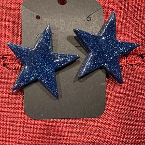 May include: A pair of blue glitter star-shaped earrings. The earrings are on a black tag with a hole for hanging.