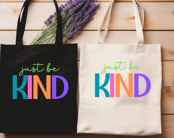 Könnte beinhalten: Zwei Tragetaschen, eine schwarze und eine cremefarbene, mit den Worten "just be KIND" in bunten Blockbuchstaben. Die schwarze Tasche hat eine grüne Schrift und die cremefarbene Tasche hat eine grüne Schrift. Zwischen den Taschen liegt ein Bund violetter Blumen.