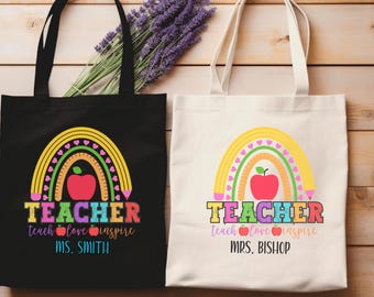 May include: Two tote bags, one black and one white, with a colorful rainbow design. The design includes a red apple, a ruler, and the word "TEACHER" in various colors. The text "teach love inspire" and a teacher's name are also included.