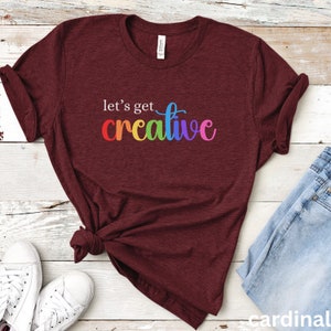 Puede incluir: Una camiseta granate con la frase "let's get creative" impresa en blanco y colores arcoíris. La camiseta está anudada a la cintura y combinada con vaqueros y zapatillas blancas. La palabra "cardinal" es visible en la esquina inferior derecha.