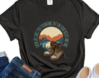 May include: Black t-shirt with a circular graphic featuring hiking boots, mountains, and the text "WHEN LIFE GIVES YOU MOUNTAINS, PUT ON YOUR BOOTS AND HIKE." The design includes shades of brown, orange, green, and blue.