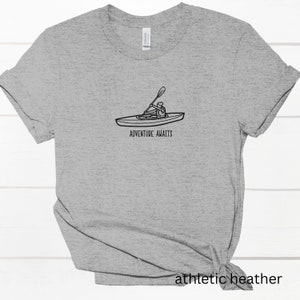 以下が含まれることがあります： グレーのTシャツに、カヤックに乗っている人の黒の線画と「Adventure Awaits」の文字が書かれています。