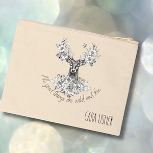 May include: A beige canvas zippered pouch with a black and white illustration of a deer's head with antlers surrounded by flowers. The text "All good things are wild and free." is printed below the image. The name "CARA USHER" is printed at the bottom of the pouch.