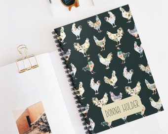 May include: A spiral-bound notebook with a dark teal cover featuring a repeating pattern of floral rooster illustrations. The notebook has the name "DONNA HOLDER" printed on a label. Other items on the white surface include sunglasses and a gold binder clip.