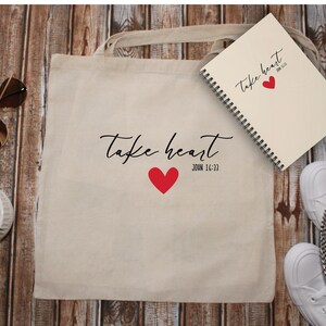 Pode incluir: Uma bolsa de lona branca com o texto "take heart" e um coração vermelho. O texto "JOHN 16:33" está abaixo do coração. Um caderno branco com o texto "take heart" e um coração vermelho está em cima da bolsa de lona.
