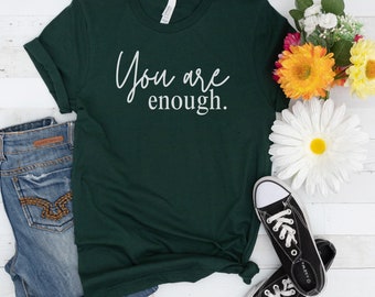Może przedstawiać: Zielony T-shirt z nadrukiem "You are enough." w kolorze białym na przodzie. Koszulka leży płasko na białym tle z parą czarnych butów z płótna i bukietem kwiatów.