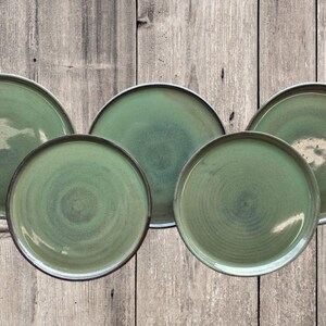 May include: Set of five green ceramic dinner plates with a subtle swirl pattern. The plates are arranged in a circle on a wooden background.