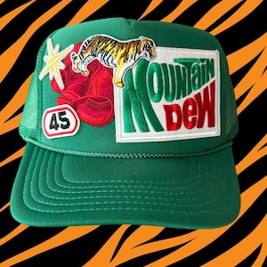 May include: Green trucker hat with a red, white, and green Mountain Dew logo patch, a tiger patch, a red flower patch, and a white star patch. The hat also has a black number 45 patch.