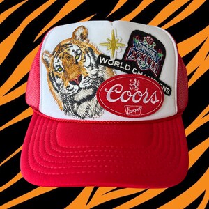 May include: A red and white trucker hat with a tiger, a star, and a Coors patch. The hat also has a Super Bowl patch that says "Super Bowl XXVII World Champions".