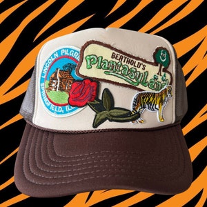 May include: A brown and beige trucker hat with a variety of embroidered patches. The patches include a red rose, a green plant, a tiger, and a patch that says "Berthold's Plantasylum".