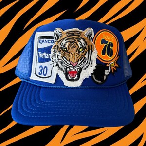 May include: Blue trucker hat with a variety of patches, including a tiger, a can of beer, a bomb, and a 76 logo.