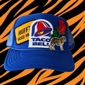 May include: Blue mesh trucker hat with various patches, including a Taco Bell logo, a tiger, a rose, and a patch that says "Ruff's Seed Farm".