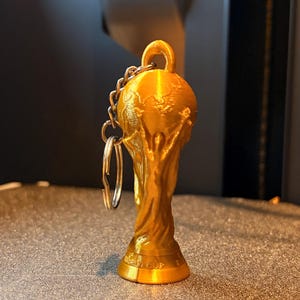 May include: A gold-colored 3D printed keychain shaped like the FIFA World Cup trophy. The object features a spherical top with a globe design, a central figure with raised arms, and a base with the embossed text WORLD CUP. A metal ring is attached.