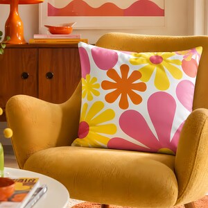 May include: A square throw pillow featuring a vibrant, repeating pattern of large stylized flowers in shades of bright yellow, hot pink, and burnt orange against a crisp white background, showcasing a bold, colorful, and graphic floral design.