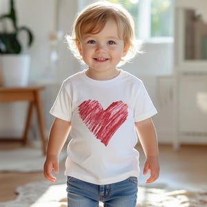 May include: A child wearing a white t-shirt with a large, red heart design. The heart appears to be hand-drawn with a scribbled texture. The child is also wearing blue jeans.