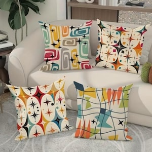 May include: Four decorative throw pillows with colorful, retro-style geometric patterns. The pillows feature designs with shapes, stars, and lines in shades of red, yellow, blue, and green on a cream background. They are arranged on a white sofa.