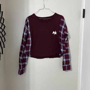 May include: A maroon cropped long-sleeve shirt with plaid sleeves. The shirt has a white logo that says "A/E" on the front.