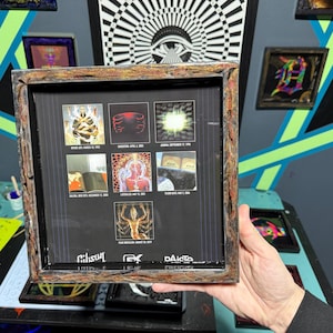 May include: A framed artwork featuring a series of images with dates and text. The frame has a textured, brown and silver appearance. The artwork is held by a person. The background includes other framed art and geometric wall art.