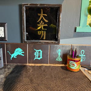 May include: A decorative kitchen scene featuring a framed black artwork with gold Japanese characters. Below, brown tiles display teal graphics. A striped container and a colorful pen holder add pops of color.