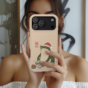 May include: A beige phone case featuring a grumpy green frog wearing a Santa hat. The frog is depicted in a Japanese art style, holding a bowl of ramen and Christmas lights. A small gift box is also present.