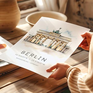 May include: A watercolor illustration of the Brandenburg Gate in Berlin, Germany. The image is on a white background with the text "BERLIN 52.5163&deg;N | 13.37825&deg;W BRANDENBURG | GERMANY" below the illustration.