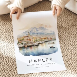以下が含まれることがあります： イタリアのナポリの水彩画で、背景にはヴェスヴィオ山が描かれています。絵は街のウォーターフロントを描いており、港にはボートが停泊しています。絵の下には大きな文字で「NAPLES」と書いてあり、その下に「40.84838&deg;N | 14.27206&deg;W」の座標と「CAMPANIA | ITALY」の場所が記されています。