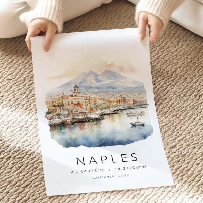 以下が含まれることがあります： イタリアのナポリの水彩画で、背景にはヴェスヴィオ山が描かれています。絵は街のウォーターフロントを描いており、港にはボートが停泊しています。絵の下には大きな文字で「NAPLES」と書いてあり、その下に「40.84838&deg;N | 14.27206&deg;W」の座標と「CAMPANIA | ITALY」の場所が記されています。