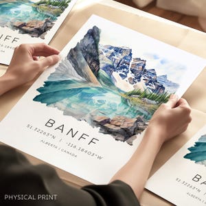 May include: A watercolor painting of a mountain lake in Banff National Park, Alberta, Canada. The painting features a turquoise lake surrounded by snow-capped mountains. The text "BANFF" is printed below the image, along with the coordinates 51.32263°N -116.18403°W and the words "ALBERTA | CANADA". The words "PHYSICAL PRINT" are printed below the coordinates.