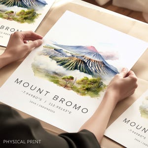 Op de afbeelding: Een aquarel-illustratie van de Mount Bromo, een vulkaan in Indonesië. De afbeelding toont een berg met een krater, omringd door wolken en groene vegetatie. De tekst "Mount Bromo -7.94980°S | 112.95143°E Java | Indonesië" is onder de afbeelding gedrukt.