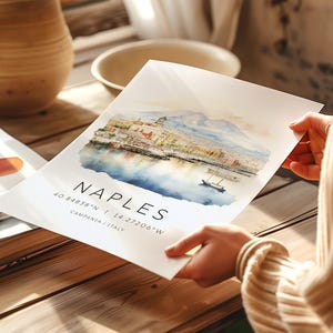 以下が含まれることがあります： イタリアのナポリ市の水彩画で、背景にはヴェスヴィオ山が描かれています。絵は白い背景に描かれており、その下に「NAPLES 40.84838&deg;N | 14.27206&deg;W CAMPANIA | ITALY」というテキストが印刷されています。