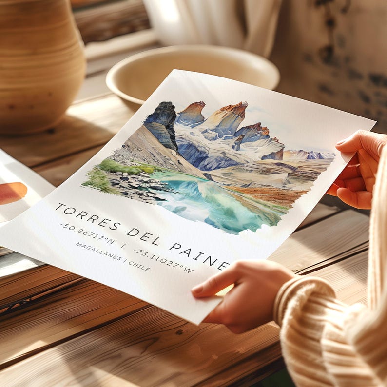 May include: A watercolor print of the Torres del Paine National Park in Chile. The print features a mountain range with a turquoise lake in the foreground. The text "Torres del Paine" is printed below the image, along with the coordinates " -50.86717&deg;N | -73.11027&deg;W" and "Magallanes | Chile".