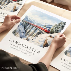 Op de afbeelding: Een aquarel illustratie van een rode trein die over een brug rijdt in een besneeuwd berglandschap. De tekst "LANDWASSER 46.68042°N | 9.67440°W GRAUBÜNDEN | ZWITSERLAND" is onder de afbeelding gedrukt. De woorden "PHYSICAL PRINT" zijn onder de tekst gedrukt.