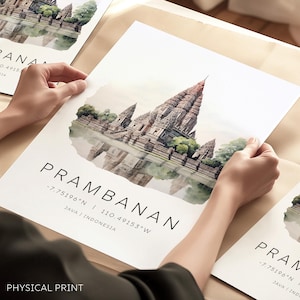 Peut inclure: Une illustration à l'aquarelle du complexe du temple de Prambanan à Java, en Indonésie. Le temple est représenté en brun clair avec un reflet dans l'eau en dessous. Le texte "PRAMBANAN -7.75196°N | 110.49153°W JAVA | INDONÉSIE" est imprimé sous l'image.