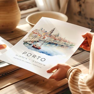 May include: A watercolor painting of the city of Porto, Portugal. The painting shows the city's waterfront with boats and buildings. The text "PORTO" is written in black at the bottom of the painting, along with the coordinates "41.15751&deg;N | -8.63706&deg;W" and the location "DISTRITO DE LISBOA | PORTUGAL".