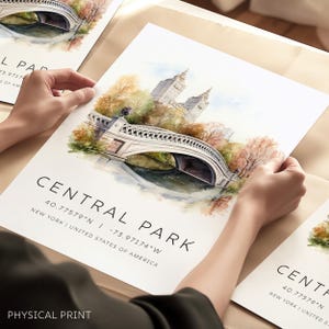 Op de afbeelding: Aquarelprint van Central Park in New York City, met de iconische Bow Bridge. Het kunstwerk toont de brug over het water, met bomen en gebouwen op de achtergrond. De print bevat de tekst "CENTRAL PARK" en coördinaten.