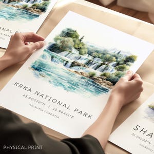 May include: A watercolor print of Krka National Park in Croatia, featuring cascading waterfalls and lush greenery. The print includes the park's coordinates and the text "PHYSICAL PRINT". The artwork is held by hands.
