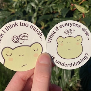 Può includere: Due adesivi rotondi con una faccia di rana verde chiaro e testo. Uno dice "Penso troppo..." e l'altro dice "E se tutti gli altri sottovalutassero?" Entrambi hanno un disegno a linea scarabocchiata sopra la rana.
