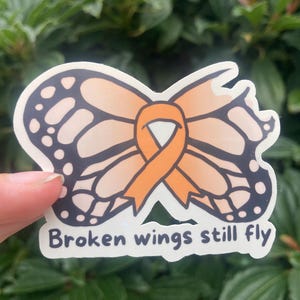 May include: A sticker featuring a butterfly design with orange and black wings, and an orange ribbon in the center. The text "Broken wings still fly" is at the bottom of the sticker.