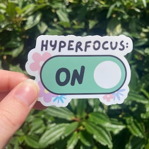Op de afbeelding: Een sticker met de tekst "HYPERFOCUS: ON" in witte letters. Het ontwerp toont een groene tuimelschakelaar met een witte cirkel, omgeven door roze en blauwe bloemaccenten. De sticker wordt tegen een achtergrond van groen gebladerte gehouden.