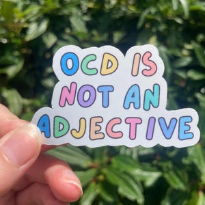 Può includere: Un adesivo bianco con la scritta "OCD IS NOT AN ADJECTIVE" in lettere colorate a bolle. L'adesivo è tenuto contro uno sfondo di fogliame verde.